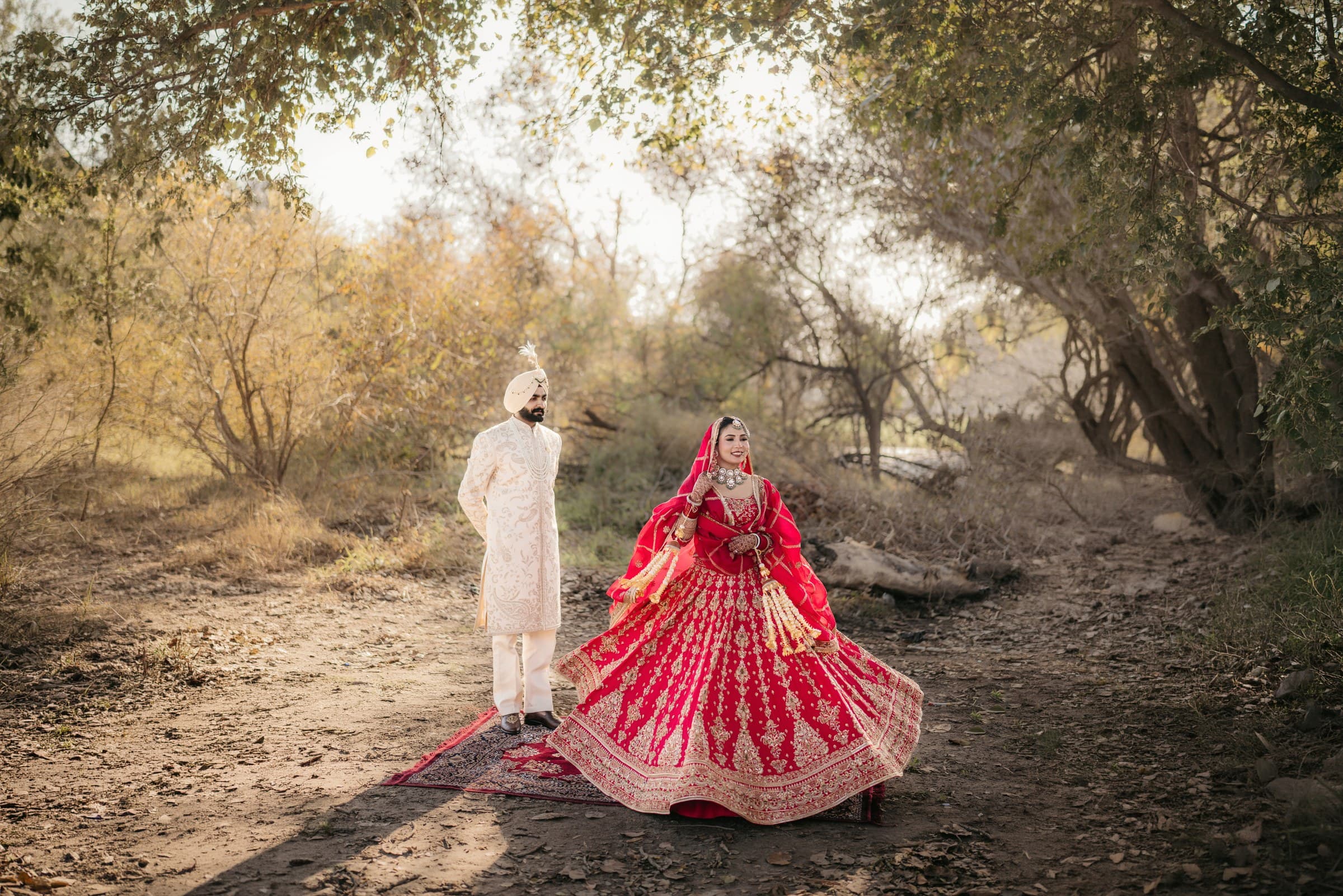 A Sikh groom in a cream sherwani watching his bride twirl in a red lehenga on a wooded path
