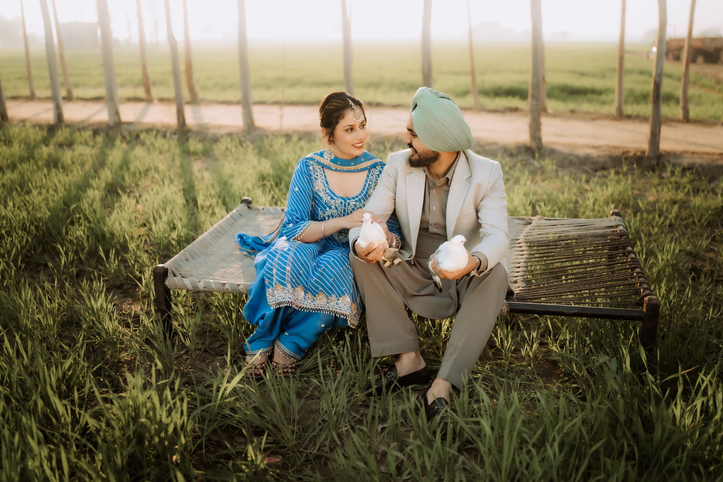 A couple sitting together on a charpai in a wheat field, each holding a white dove