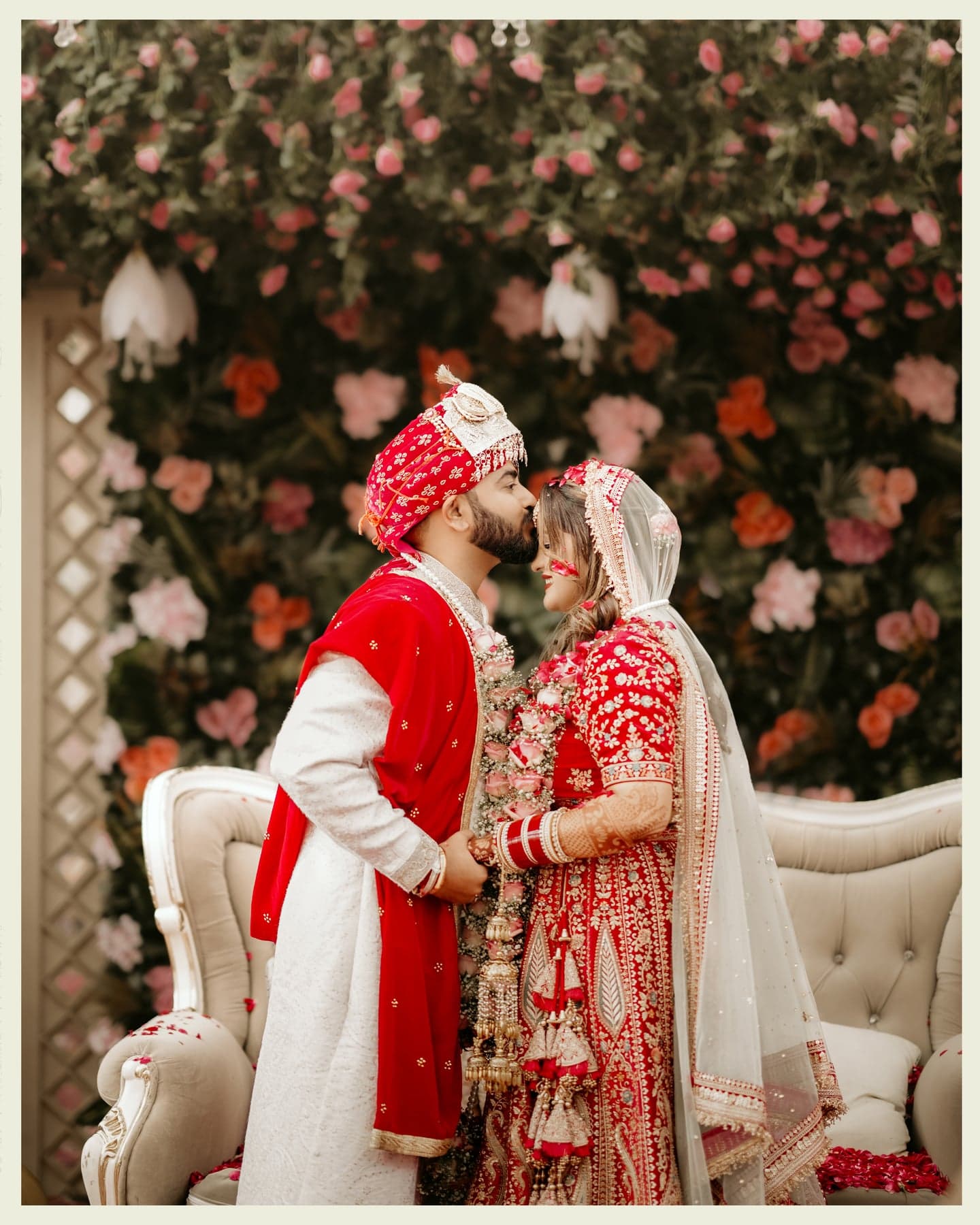 A groom kissing a bride's forehead in front of a wall of roses